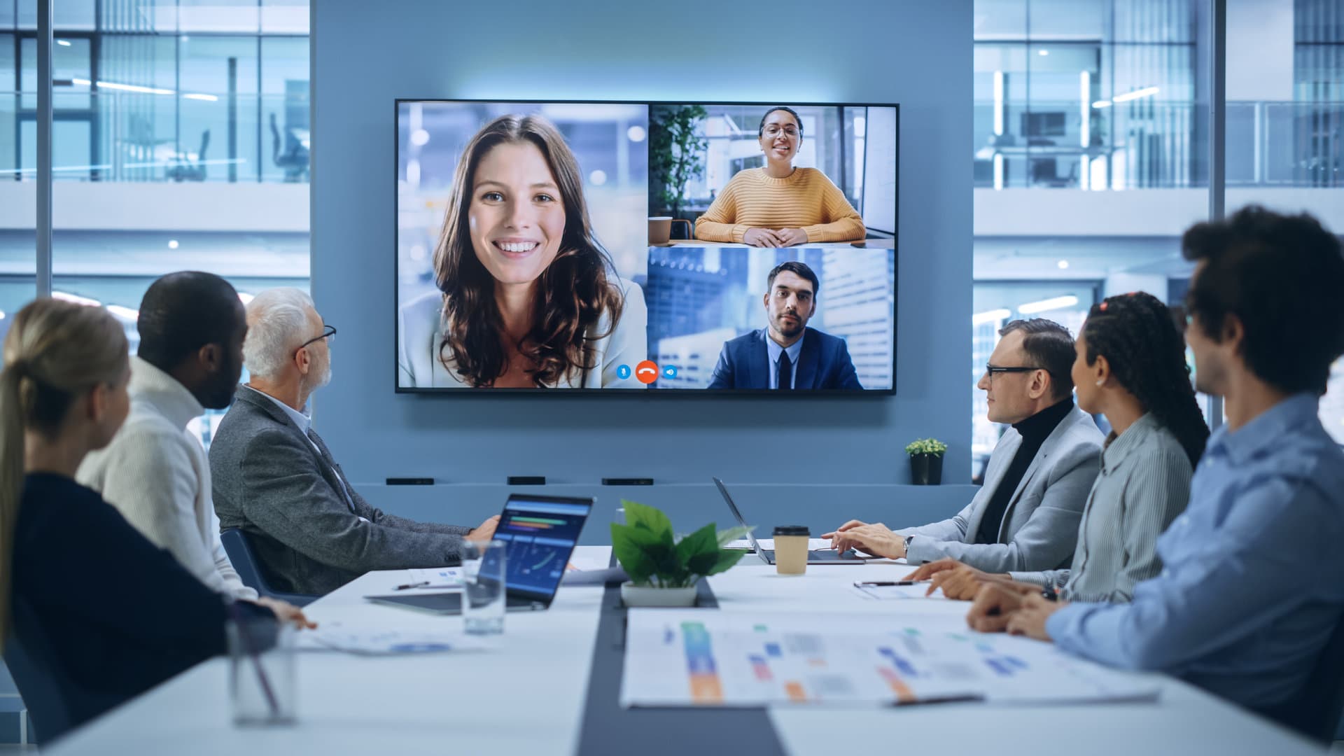 An executive team in a boardroom on a video call with three remote colleagues